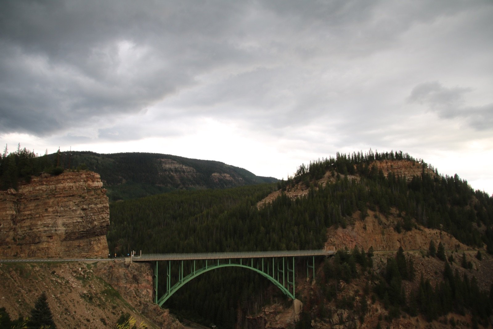A green steel bridge arching across a forested canyon under a stormy sky